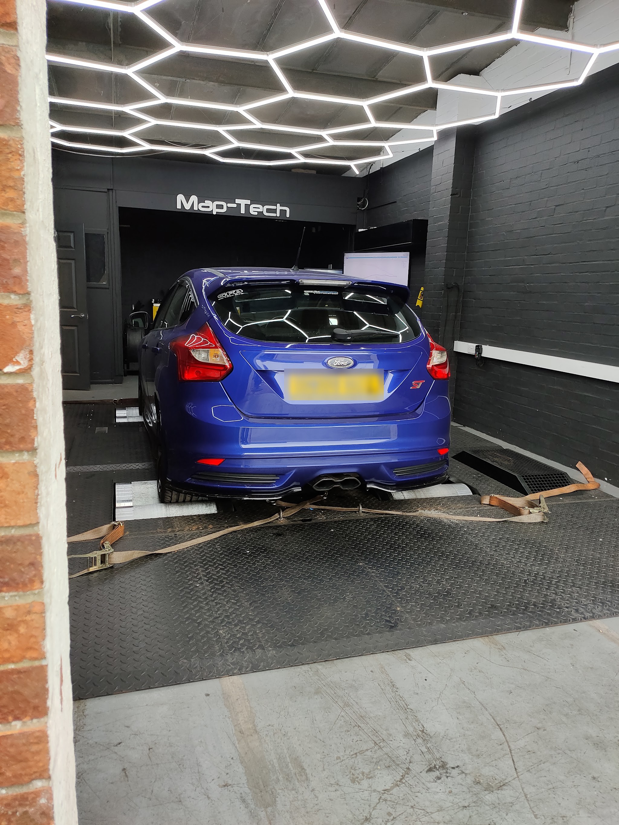 A blue Ford Focus ST strapped on the rollers in the Map-Tech dyno cell under the hexagon LED ceiling